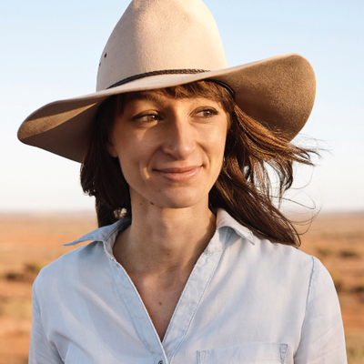Anika is smiling, she has shoulder length brouwn hair and she is wearing a wide brim hat and has red dirt around her