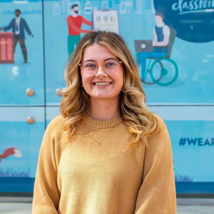 Claudia is smiling with shoulder length blonde hair, a brown knitted jumper and glasses.